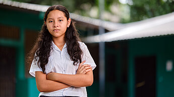 Mädchen steht mit verschränkten Armen vor einer Schule.